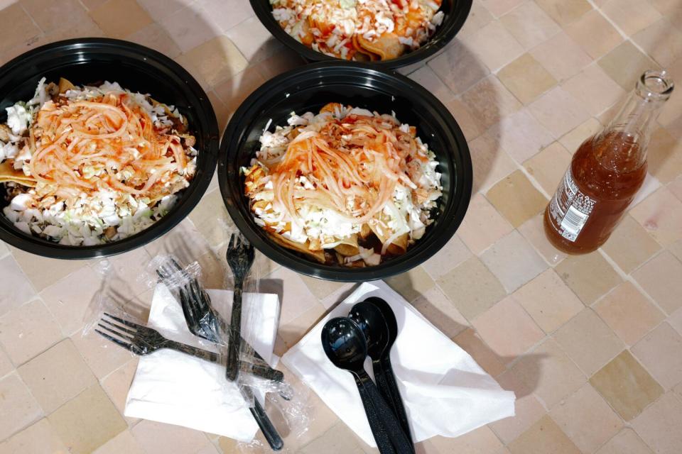 Three black plastic bowls of tacos, with an open bottle of soda on the right and sets of plastic utensils and napkins.