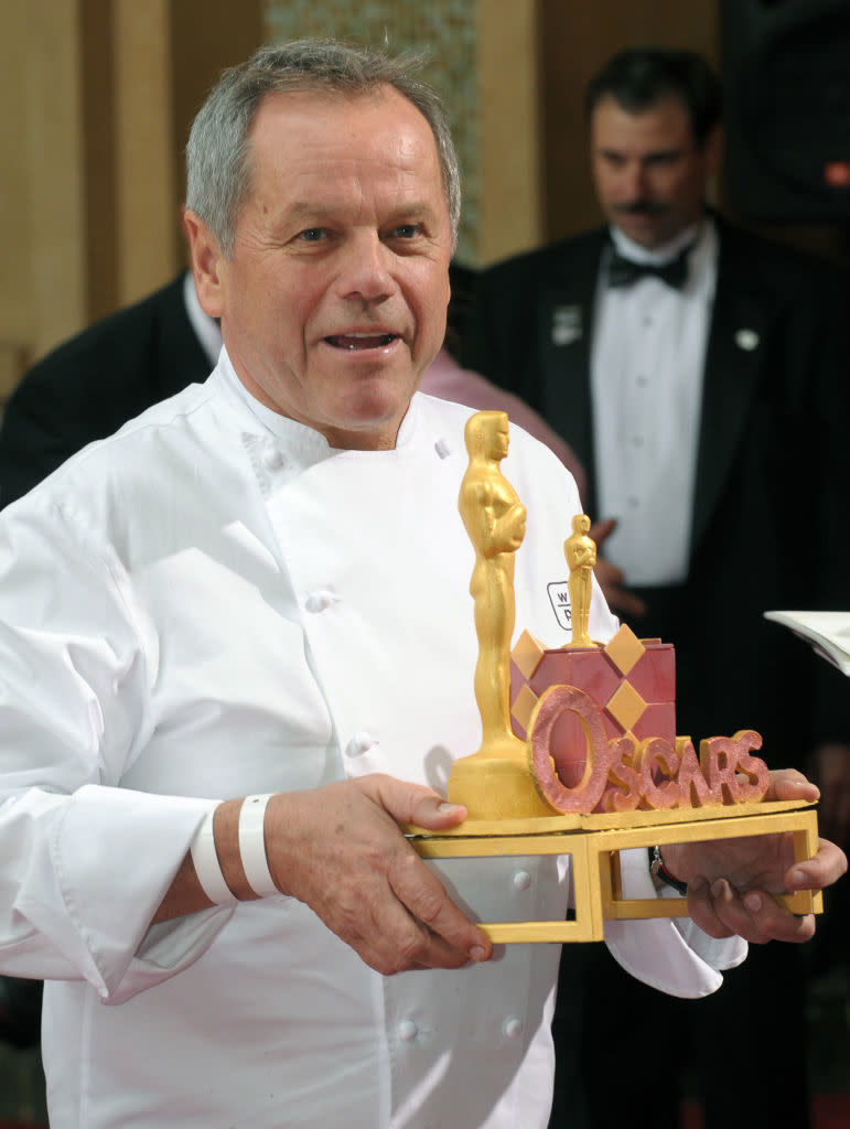 Wolfgang Puck holding an Oscars-themed chocolate creation, wearing a white chef's coat. A man in a tuxedo stands in the background