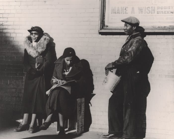 A vintage black and white image show two Black women and one Black man in the sunshine before a brick wall