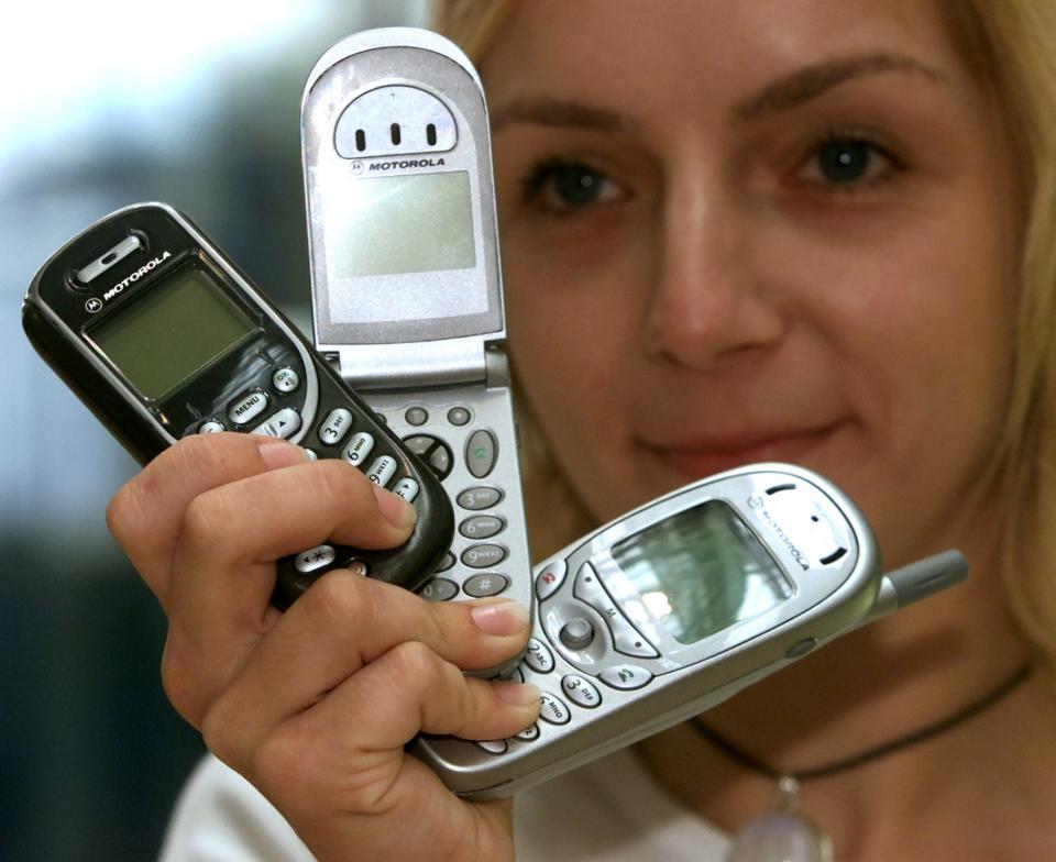 a blonde woman holds up three motorola phones, the Talkabout 192, v66, and Timeport 280
