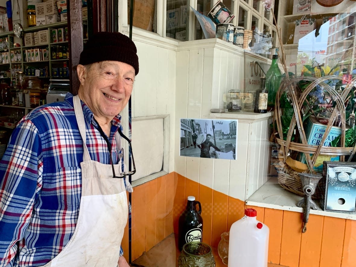 Angelo Tosi, 90, stands outside the imported Italian food shop on Main Street, Vancouver he inherited from his father. In the black-and-white photo to his left is him as a young man carrying flour into the store, which was previously located on nearby Union Street in Strathcona neighbourhood. (David P. Ball/CBC - image credit)