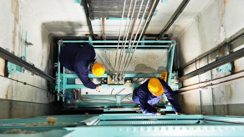 two male technician machinist worker at work adjusting elevator mechanism of lift with spanner.