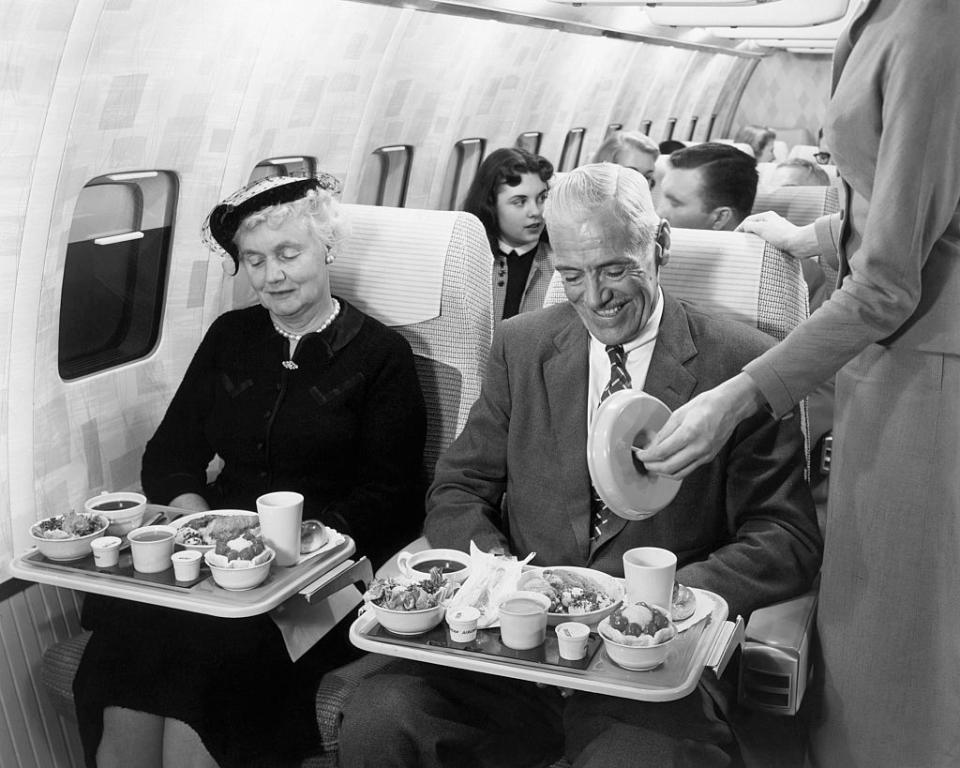 An elderly woman and man are seated in an airplane, receiving meal service from a flight attendant. Other passengers are visible in the background