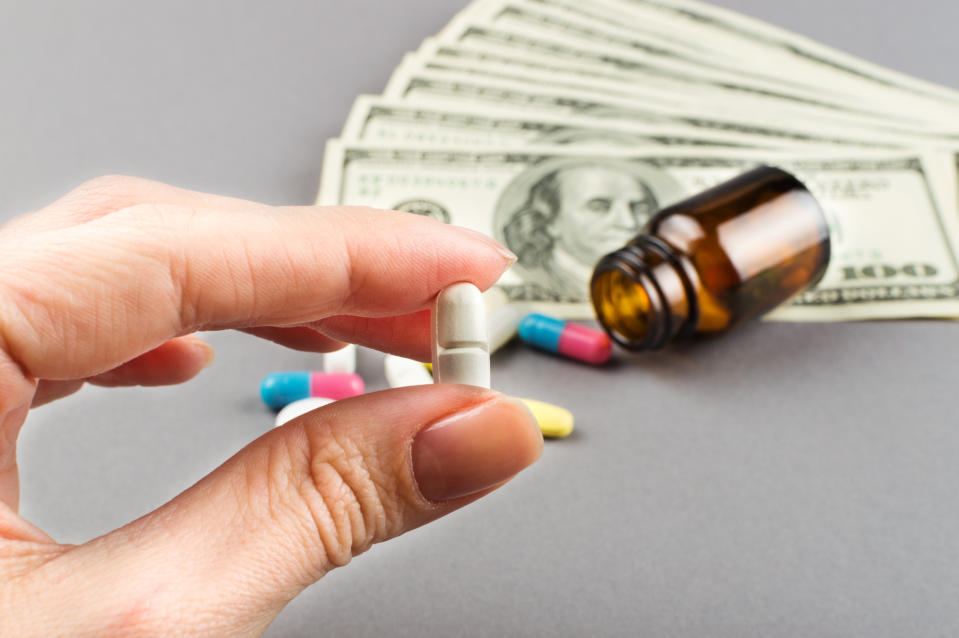 Hand holding a pill with $100 bills, a pill bottle and other pills on a surface in the backgorund