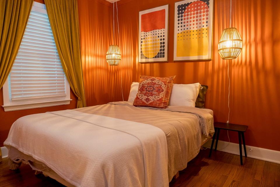 The primary bedroom continues the eclectic feel of the house with orange walls and complimentary-colored art on the walls. Basket lights provide a decorative touch.