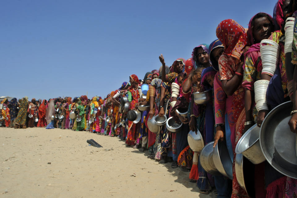 Women with colorful head coverings and bangles carry huge metal pails as they wait in a long line on desert ground.