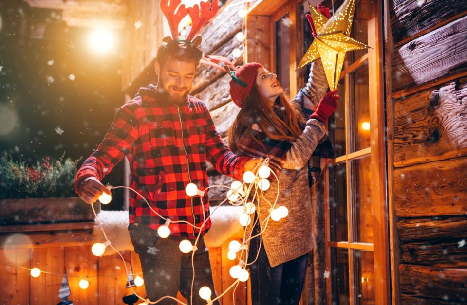 Man wearing buffalo check shirt and woman with a beret  hanging holiday lights and star decor outside.