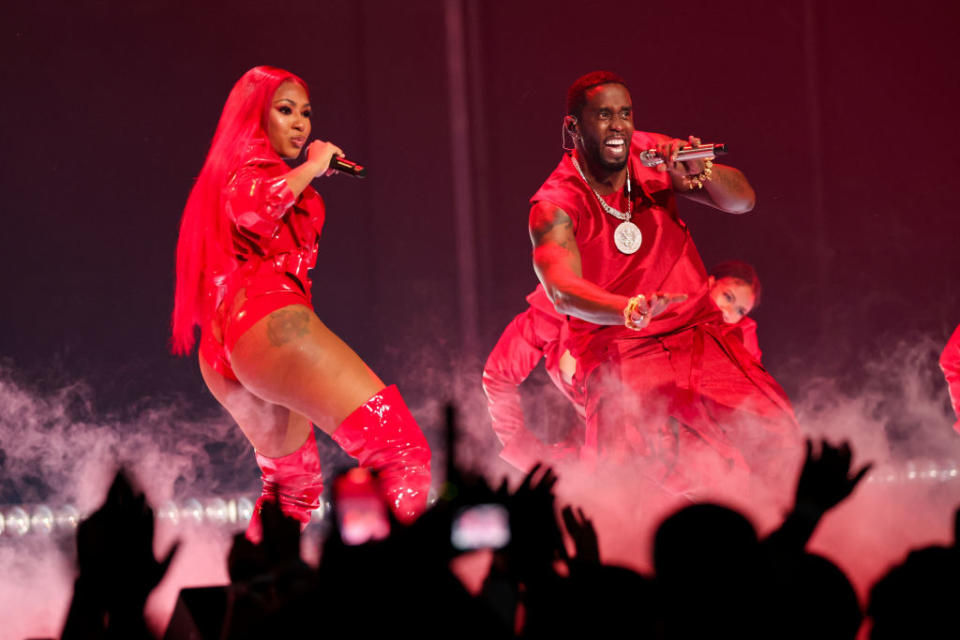 Yung Miami and Diddy perform on stage wearing stylish red outfits, with fog effects and audience hands raised in the foreground