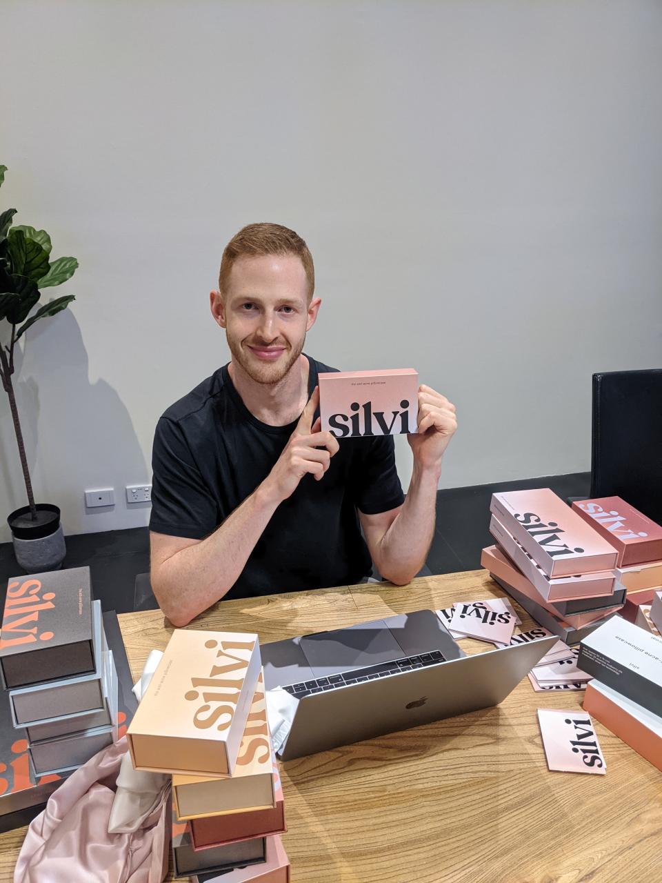 Max Hertan sits a table with his laptop and boxes of Silvi pillows, holding one pink box up. He wears a black T-shirt and has short cropped hair.