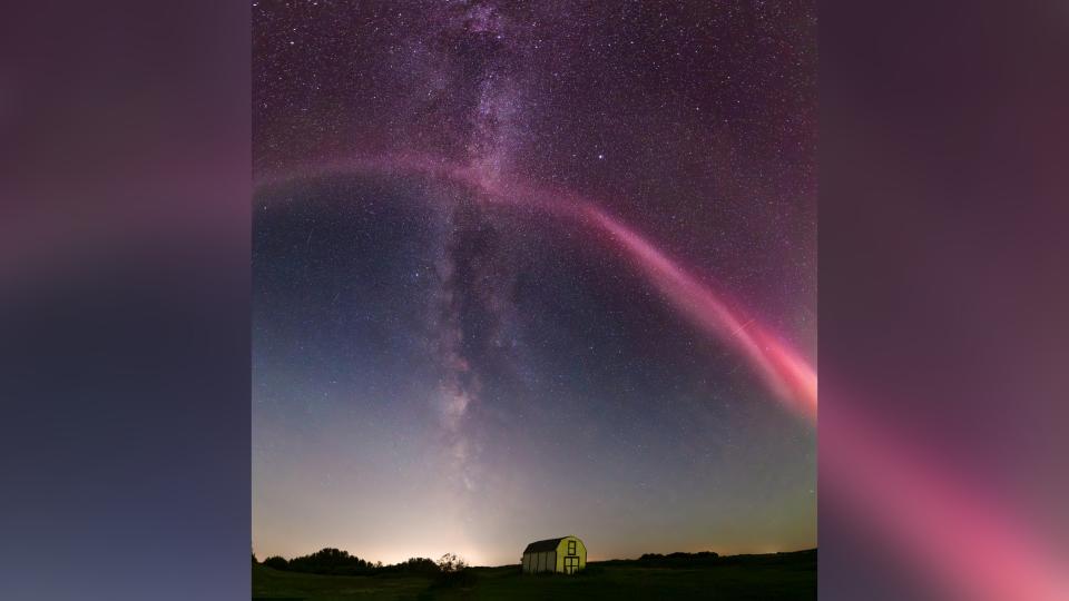  A long red stream of light arches over the night sky, in the background is the milky way appearing as a thick band of stars running from the top of the image down to the horizon. 