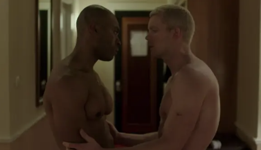 Two shirtless men, one with a dark complexion and the other with a light complexion, face each other closely in a hallway, sharing an intimate moment. Names unknown