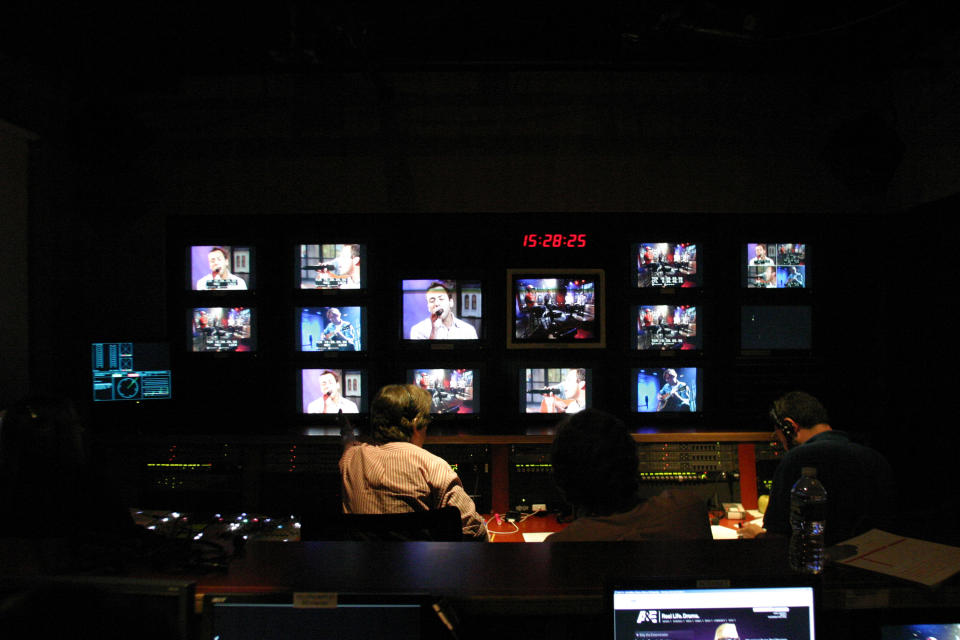 people sitting in a broadcast control room watching multiple televisions