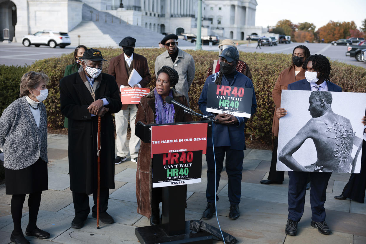 Rep. Sheila Jackson Lee at the microphone at a podium marked: HR40 Can't Wait, flanked by someone holding a poster of a black-and-white historical photo showing an African-American man looking away from the camera to display the horrific scars from whippings on his back.