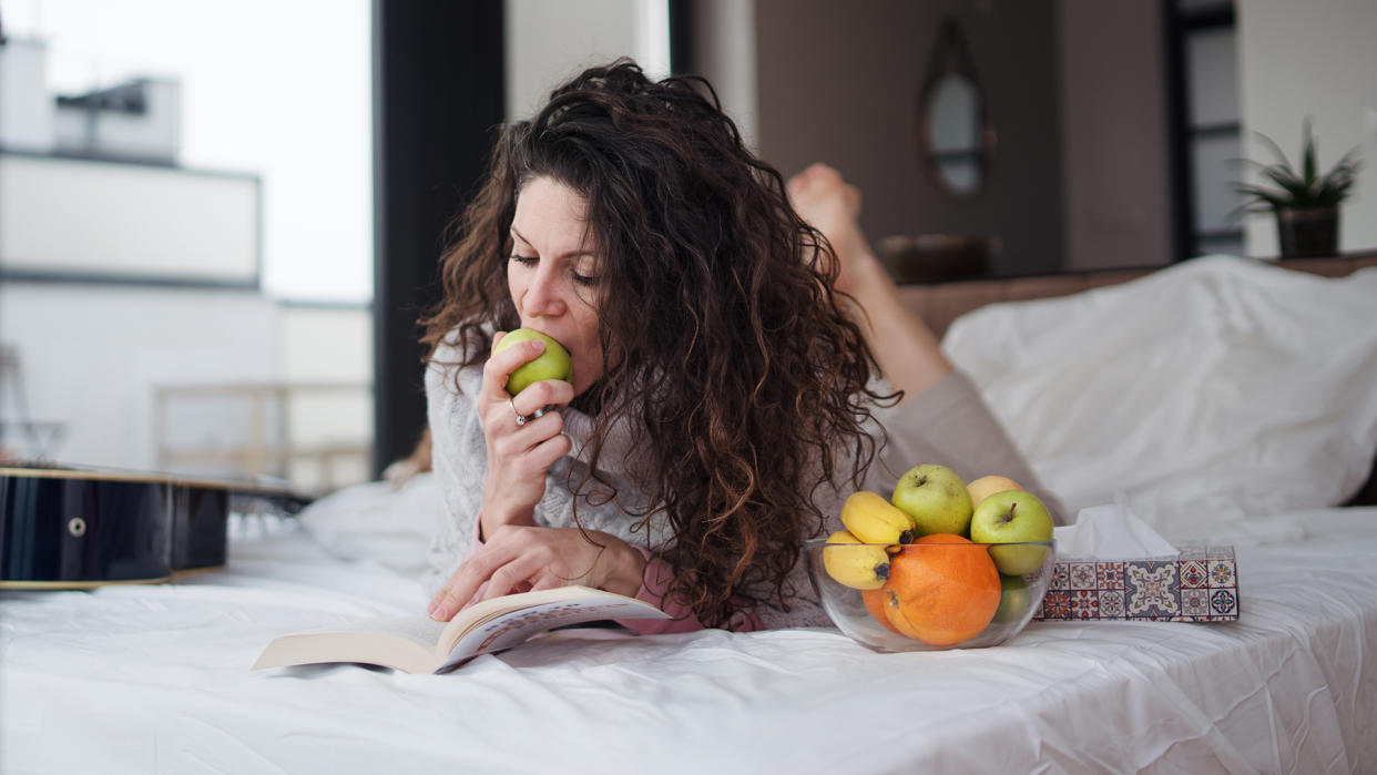  A woman lying on a bed eating fruit. 