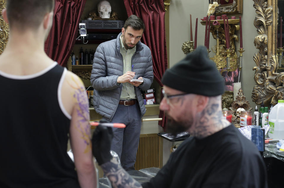 In this Thursday, April 11, 2019 photo Berkshire Eagle writer Benjamin Cassidy, behind center, takes notes as tattoo artist Brian Brown, of Dalton, Mass., right, creates an image on the arm and shoulder of customer Scott Forrest, of North Adams, Mass., left, at a tattoo parlor in Dalton, Mass. The paper now features a new 12-page lifestyle section for Sunday editions, a reconstituted editorial board, a new monthly magazine, and the newspaper print edition is wider. That level of expansion is stunning in an era where U.S. newspaper newsroom employment has shrunk by nearly half over the past 15 years. (AP Photo/Steven Senne)