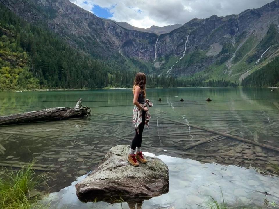 Emily stands on a rock in the middle of a lake with clear water in Glacier National Park. The lake is surrounded by mountains and trees.