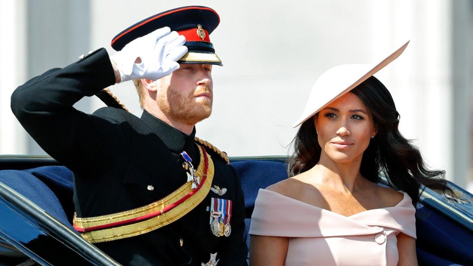 Prince Harry saluting in uniform as he sits in a carriage with Meghan Markle wearing a pink off shoulder dress with a matching hat.