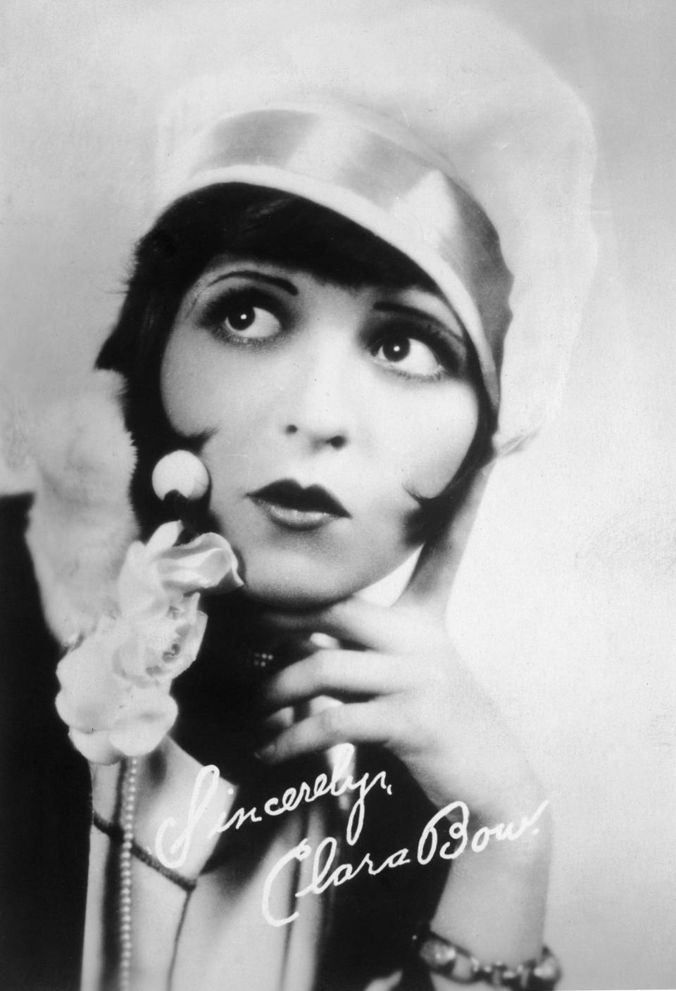 Autographed studio portrait of American actor Clara Bow (1905-1965) wearing a cloche hat and a bobbed hairstyle while holding her hand to her face and pursing her lips.