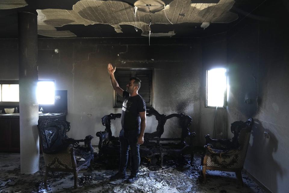 A man wearing black clothes reaches his hands up as he stands inside a charred room with two windows.