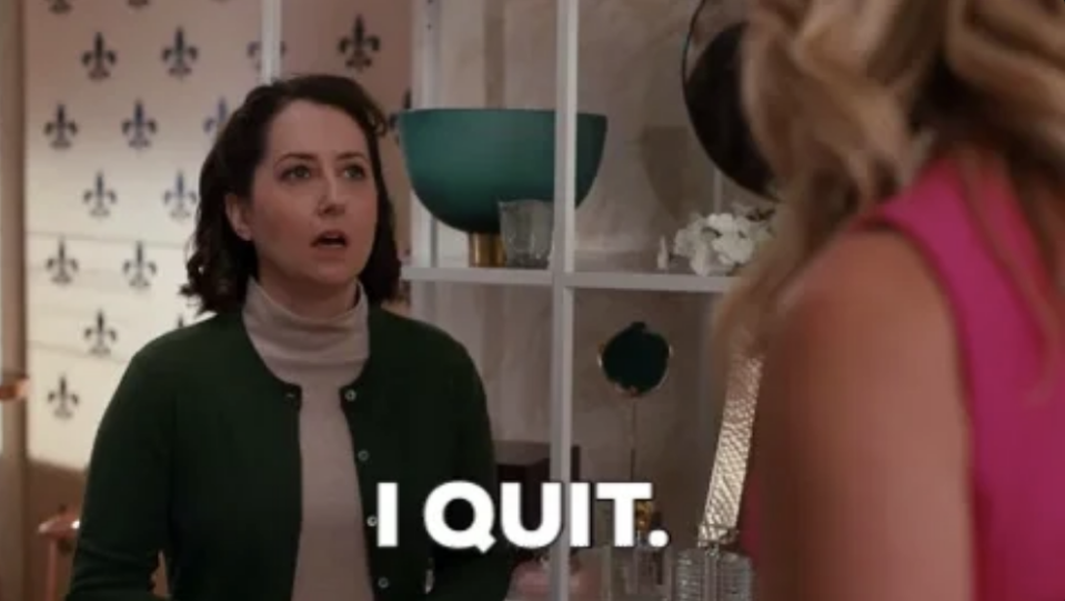 A woman in a green cardigan and beige turtleneck says "I QUIT" while talking to another woman in a pink outfit