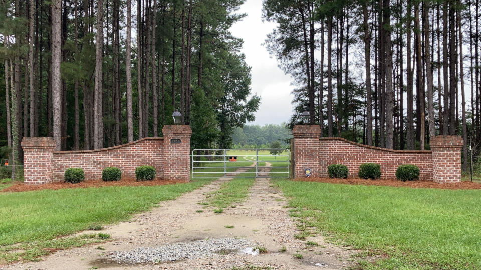 A brick-framed gate with a "no trespassing" sign in front of a road leading into a meadow, surrounded by tall, spindly trees