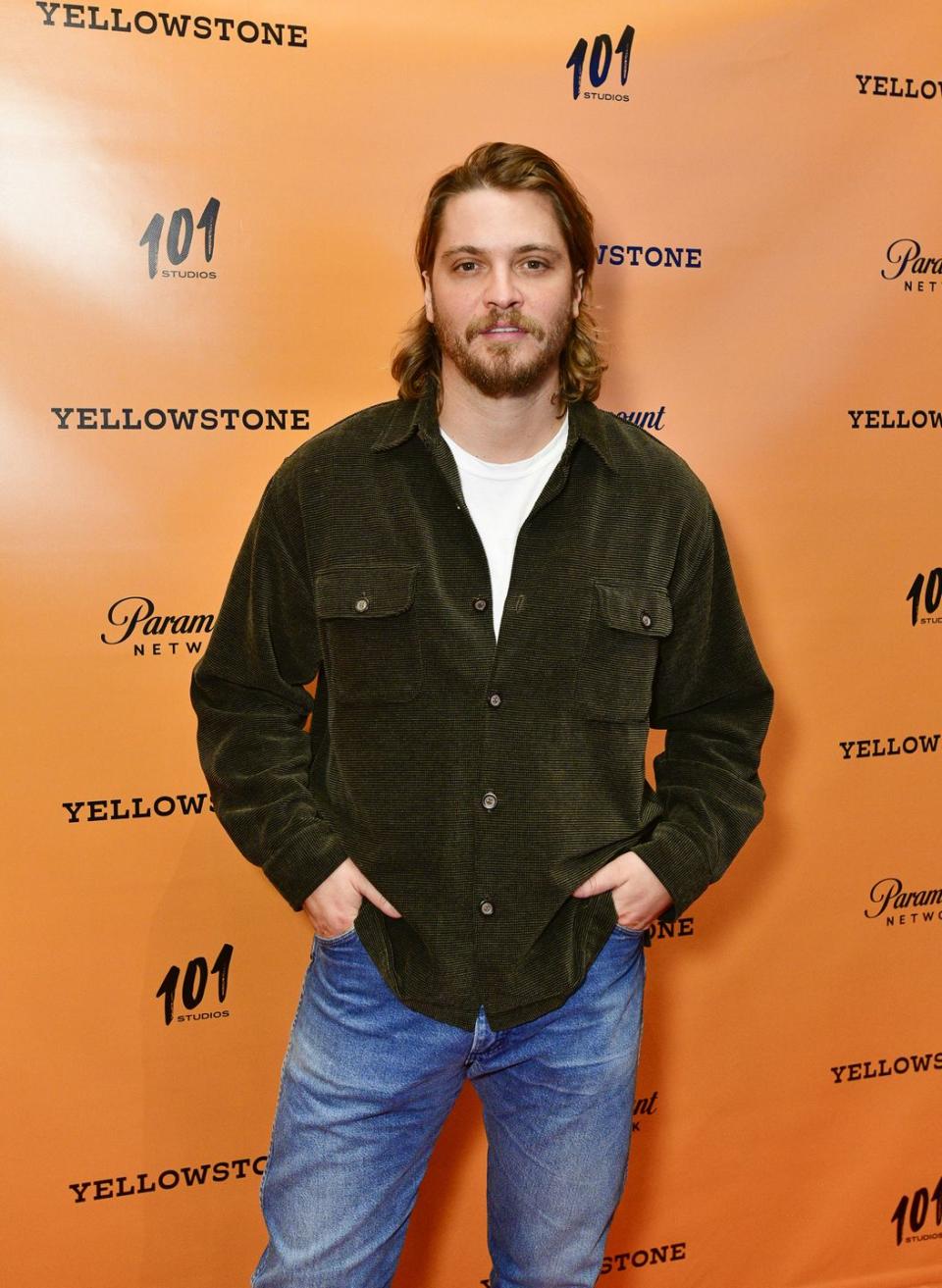 luke grimes wears his hair down to his shoulders and sports a corduroy shirt and white t shirt