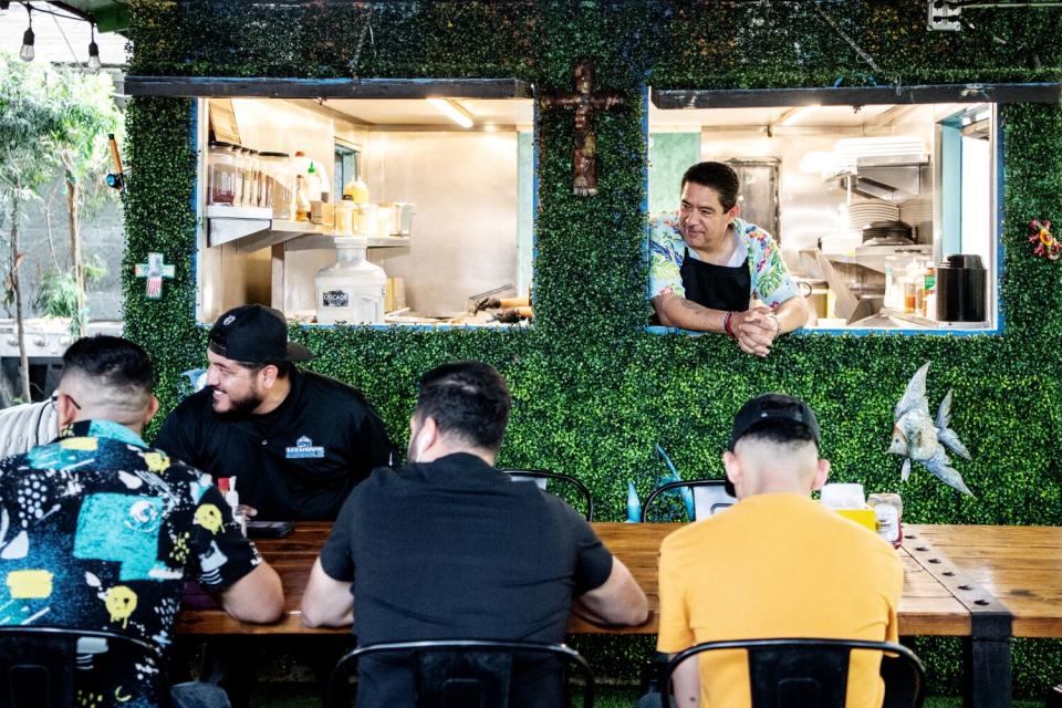 A man looks out through a window at people dining outdoors.