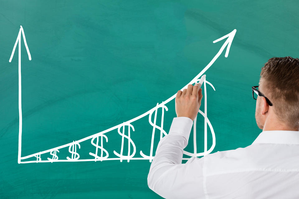 A man drawing a rising chart of dollar signs on a green chalkboard.