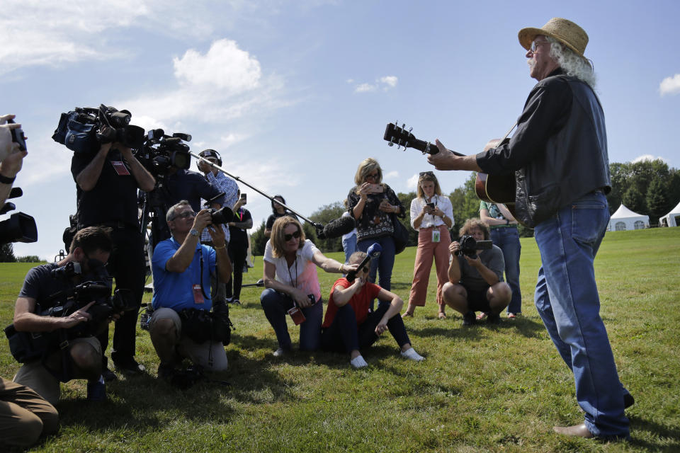 Members of the media record as Woodstock veteran Arlo Guthrie plays a song at the original site of the 1969 Woodstock Music and Arts Fair in Bethel, N.Y., Thursday, Aug. 15, 2019. Guthrie is schedule to play a set on the top of hill nearby but told reporters he wanted to play at least one song on the original 1969 site. Woodstock fans are expected to get back to the garden to mark the 50th anniversary of the generation-defining festival. Bethel Woods Center for the Arts is hosting a series of events Thursday through Sunday at the bucolic 1969 concert site, 80 miles (130 kilometers) northwest of New York City. (AP Photo/Seth Wenig)