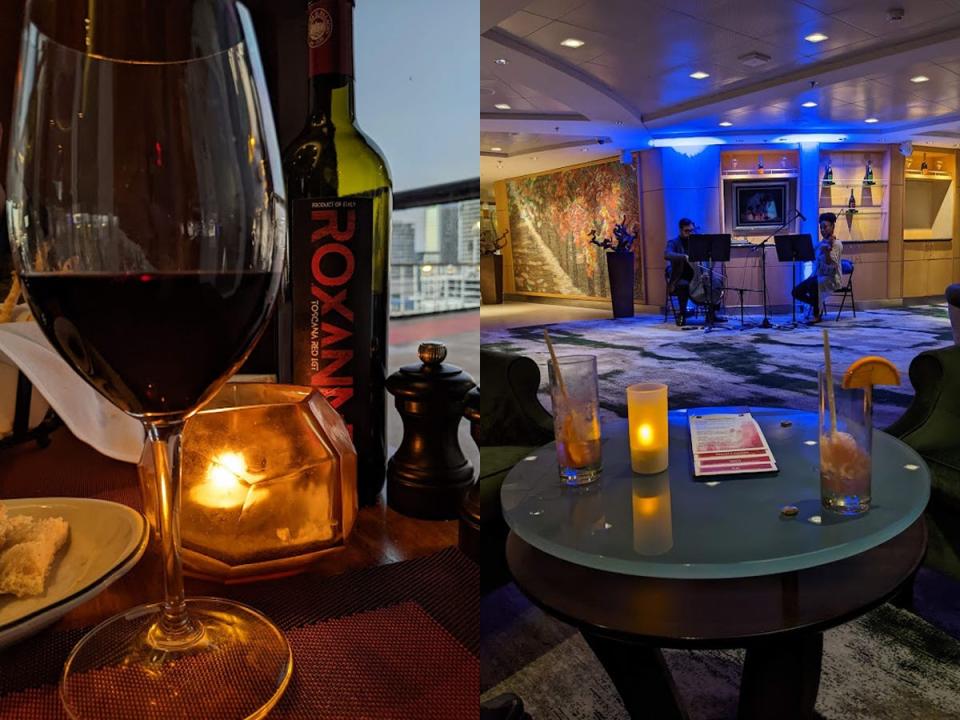 A glass of red wine and a small candle on a table next to a shot of a string quartet playing at one end of a large dining room.