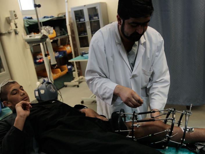 Un hombre acostado boca arriba mira un artilugio de metal envuelto alrededor de su pierna mientras un hombre con una bata blanca examina el dispositivo.