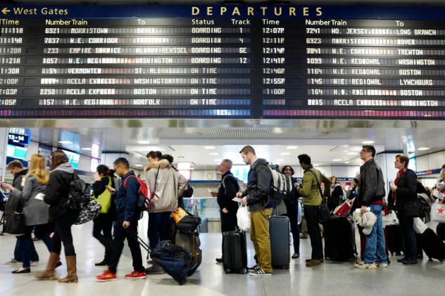 Passengers lugging suitcases and backpacks stand in a line beneath an electronic signboard in New York's Penn Station as they wait to board a train.