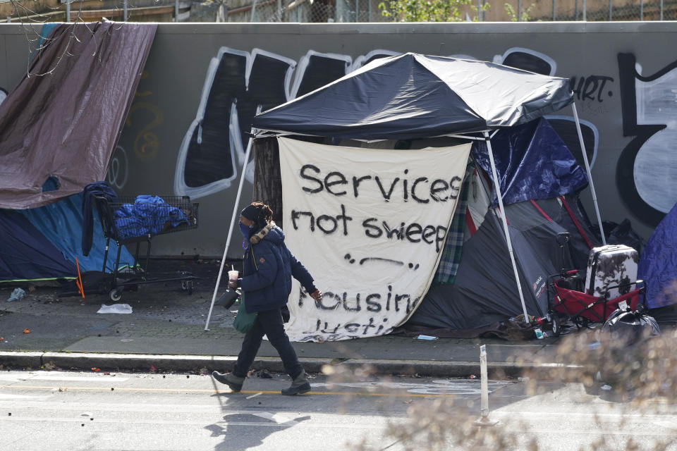 A person walks past a tent used by people experiencing homelessness with a sign on it that reads "services not sweeps," Tuesday, March 1, 2022, in downtown Seattle, across the street from City Hall. For years, liberal cities in the U.S have tolerated people living in tents in parks and public spaces, but increasingly leaders in places like Portland, Oregon, New York and Seattle are removing encampments and pushing other strict measures that would've been unheard of a few years ago. (AP Photo/Ted S. Warren)