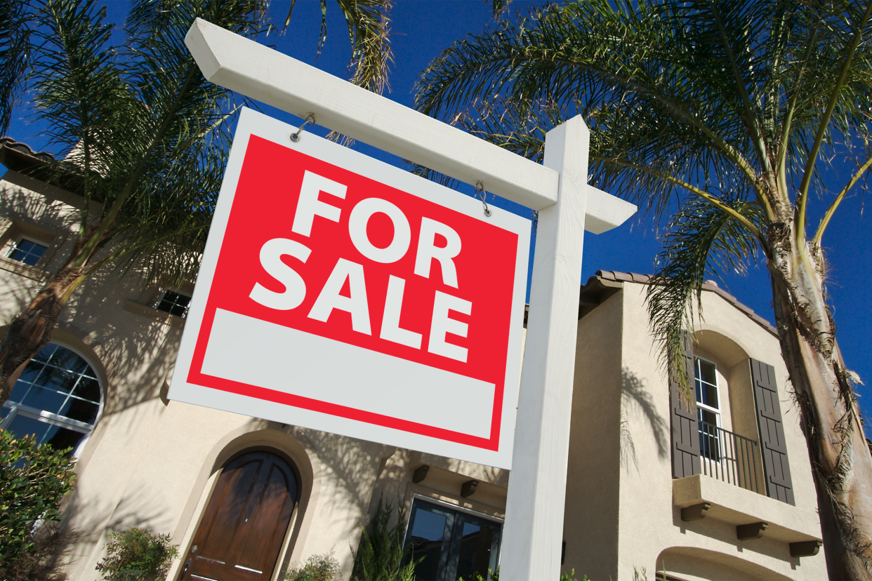 Closeup of 'For Sale' sign with white sign holder in yard of home with closeup of palm trees and home in the background