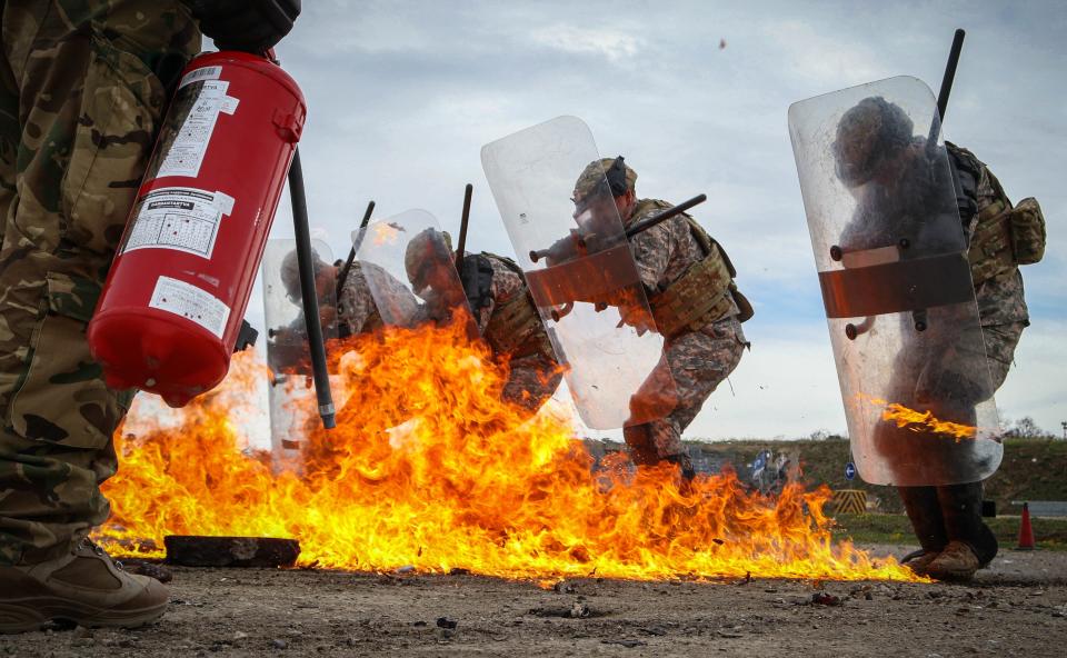 A photo of a soldier holding a fire extinguisher in front of soldiers with riot shields standing near fire.