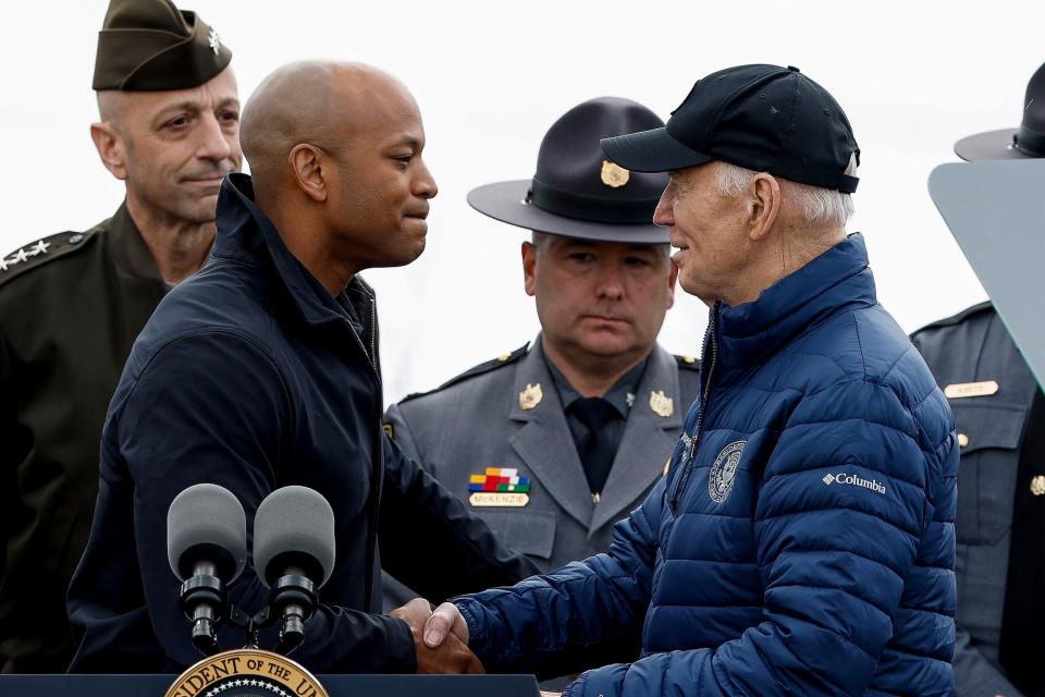 Maryland Gov. Wes Moore shakes hands with President Joe Biden behind a podium, with military and law-enforcement personnel standing behind them.