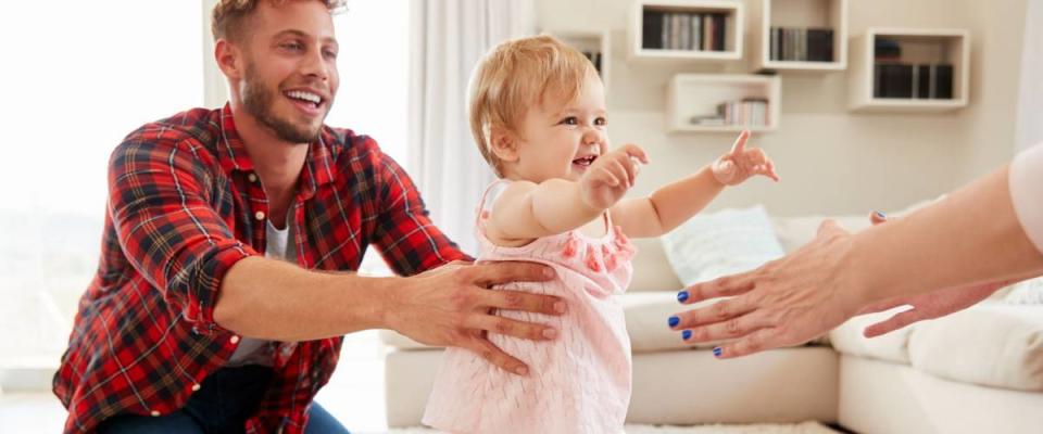 Toddler girl walking from dad to mumÕs arms in sitting room