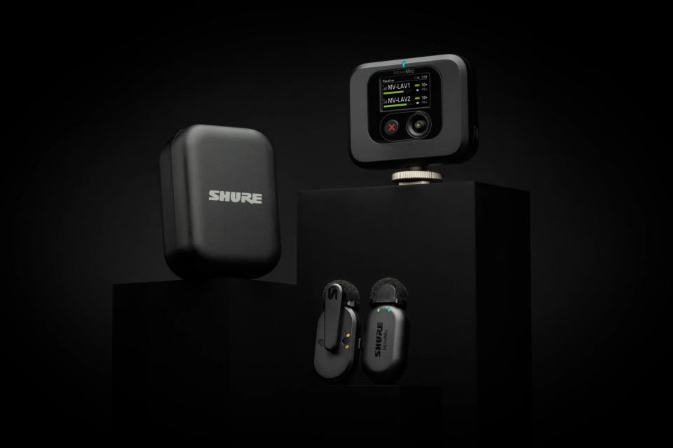 Product photo of the Shure MoveMic Two set.  Two wireless lavalier microphones, charging case and receiver sit on black pedestals in front of a dramatic black backdrop.