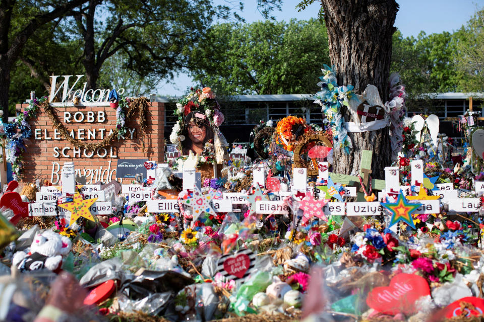 Bouquets of flowers and markers with the names of the children killed are stacked in front of sign saying: Welcome Robb Elementary School, Benvenida.