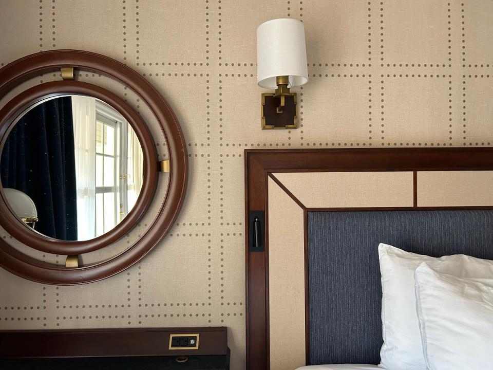 Round mirror with wood edge and corner of wood headboard with small wall sconce above