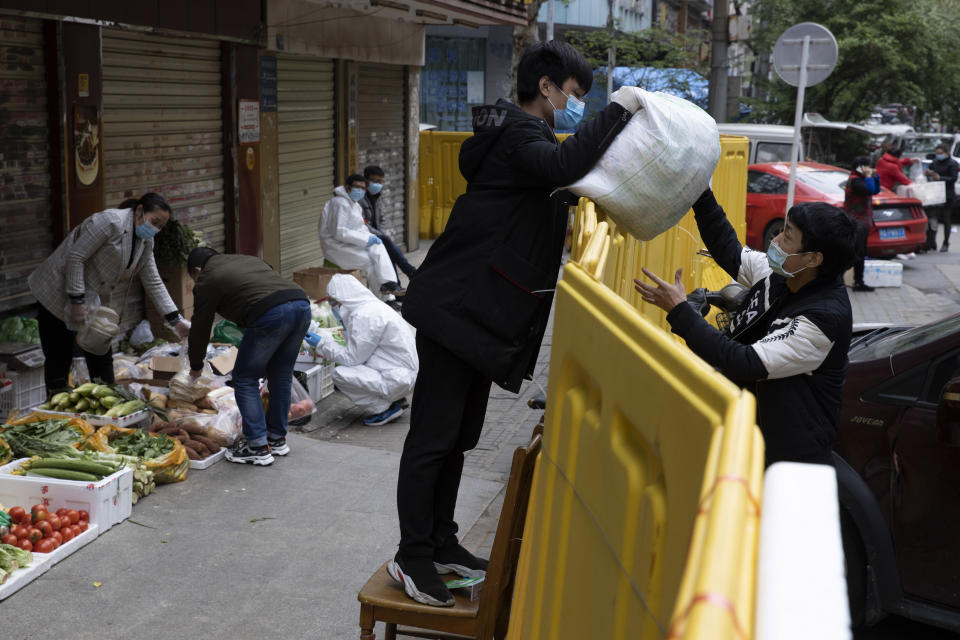 A vendor hands over grocery to another across barriers used to seal off a neighborhood to help curb the spread of the coronavirus in Wuhan, China, Friday, April 3, 2020. Sidewalk vendors wearing face masks and gloves sold pork, tomatoes, carrots and other vegetables to shoppers Friday in the Chinese city where the coronavirus pandemic began as workers prepared for a national memorial this weekend for health workers and others who died in the outbreak. (AP Photo/Ng Han Guan)