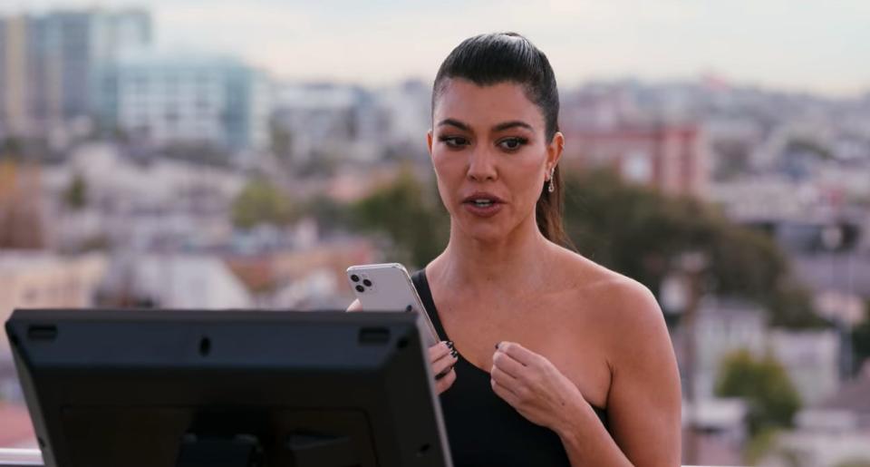 Kourtney Kardashian in "He's All That" sitting on a spin bike outside and holding a phone .