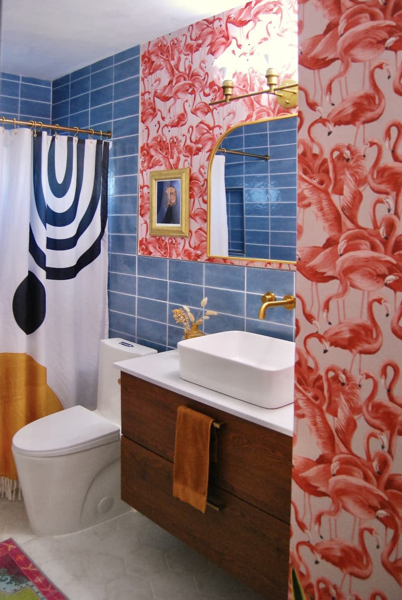 Bathroom with blue tole walls and one flamingo wallpaper accent wall, gold hardware and decor accents, wood sink cabinet with basin sink, shower curtain with bold graphic design in ochre, white, black