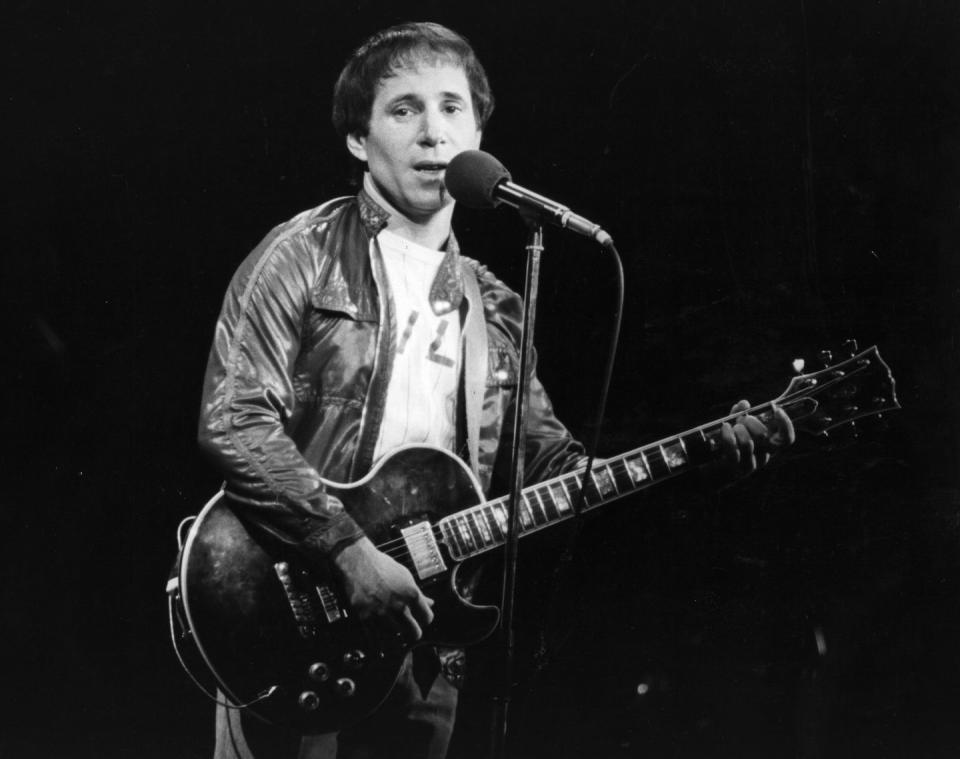 paul simon sings into a microphone on a stand while he plays electric guitar