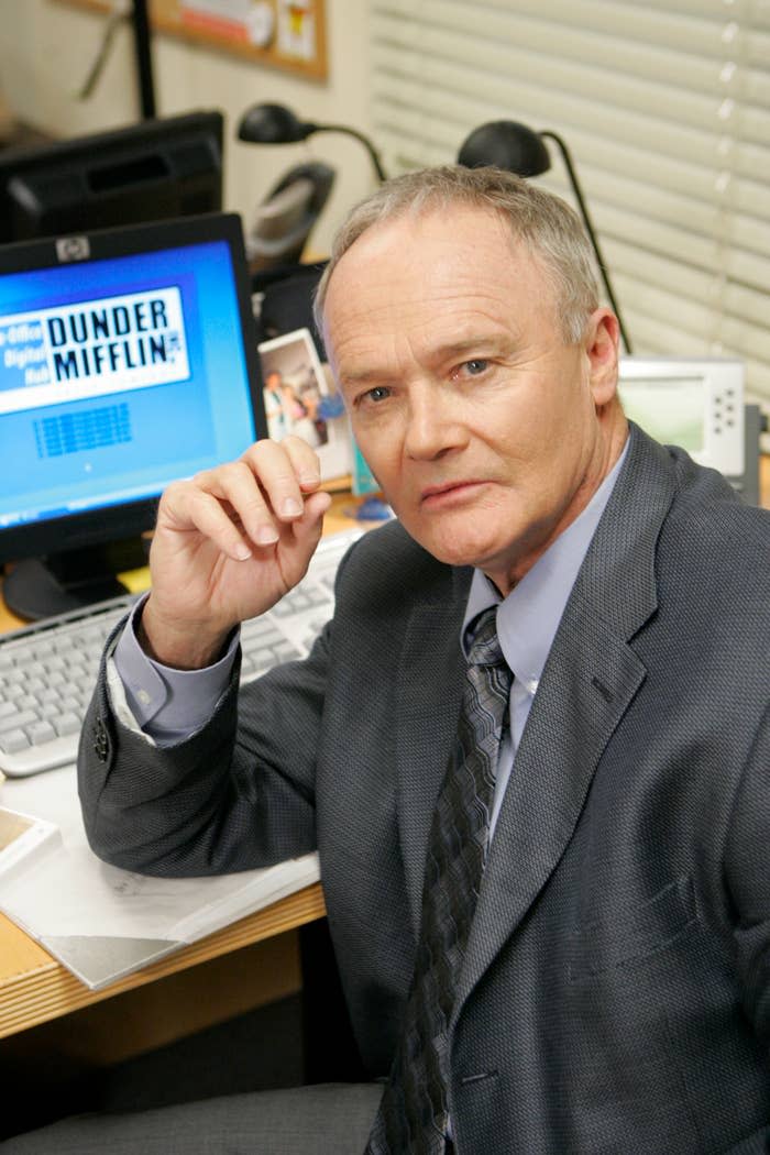 Creed Bratton, in a suit and tie, sits at a desk with a computer displaying "Dunder Mifflin" in the background