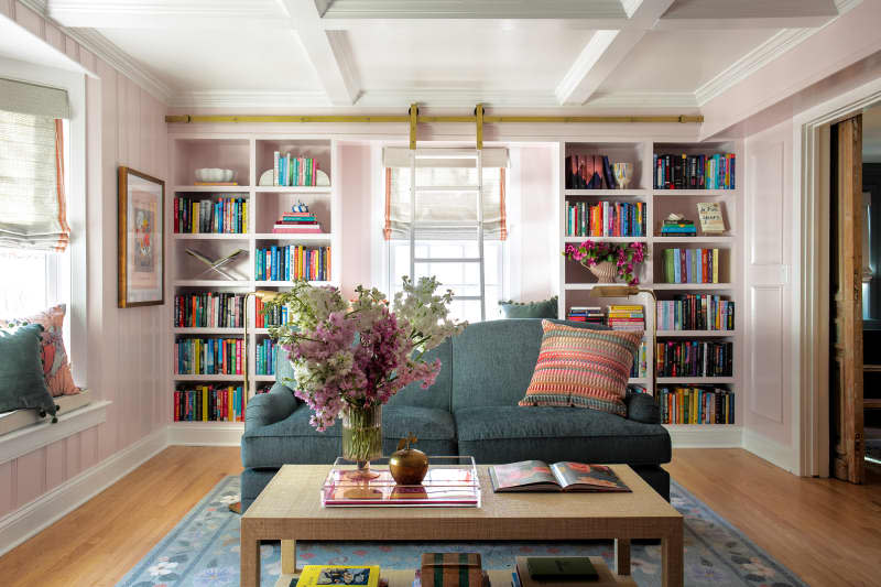 Teal loveseat with multicolored throw pillow and wood coffee table with flowers in light pink library with built-in shelves.