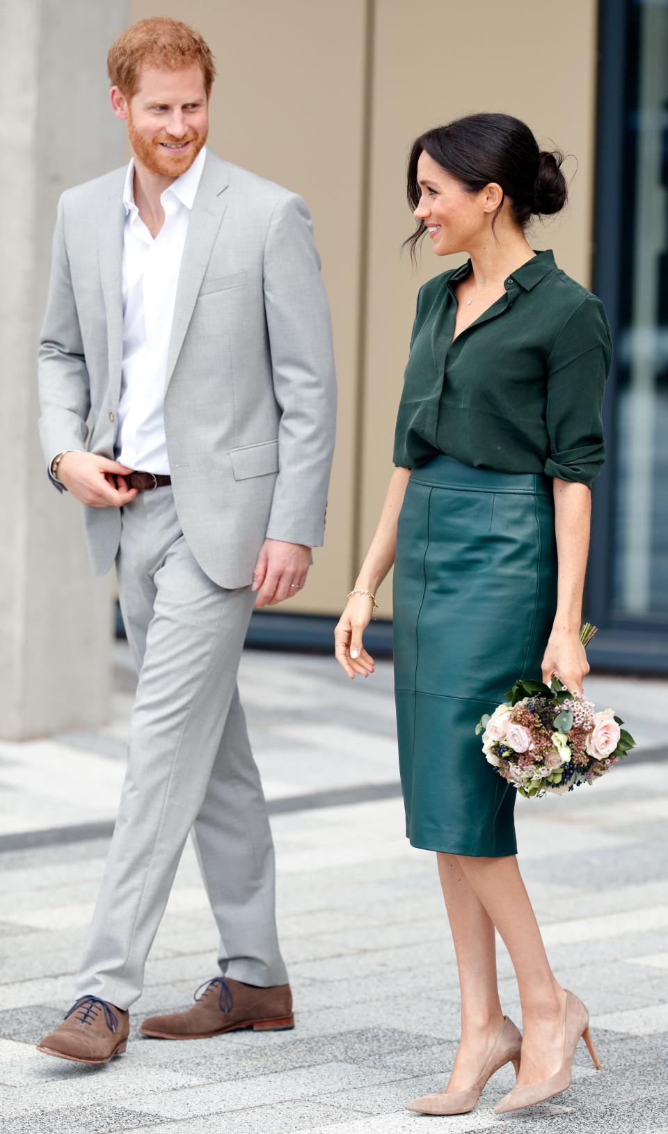 Harry in a light grey suit and button down shirt and Meghan in a dark green blouse and dark green leather skirt.