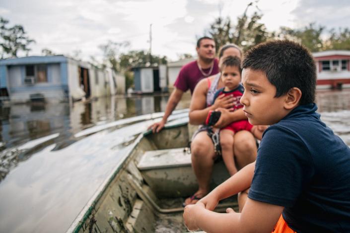 A boy with a furrowed brow and sad look sits in a boat with his family as they check on flooded mobile homes.
