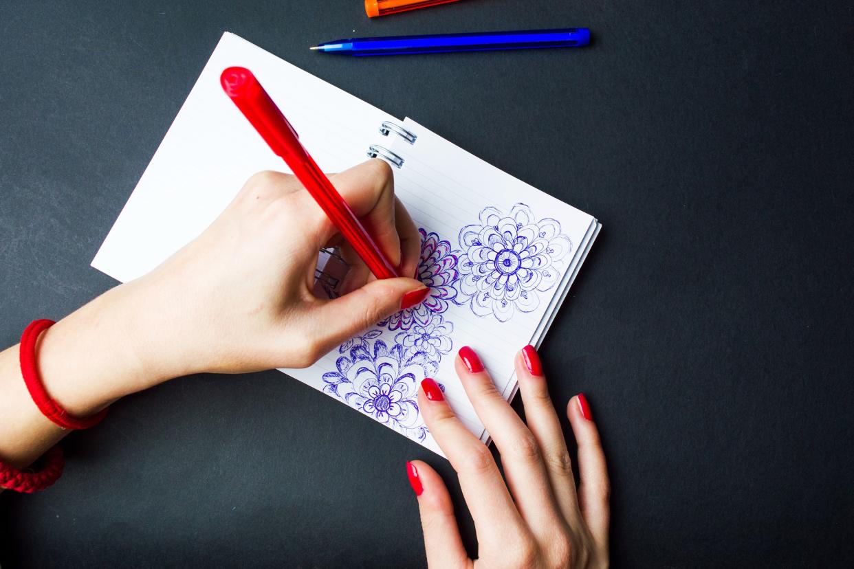Female left hand holding a red pen with blue ink, doodling flowers on a blank sheet journal with the right hand holding it on a black desk with a blue pen