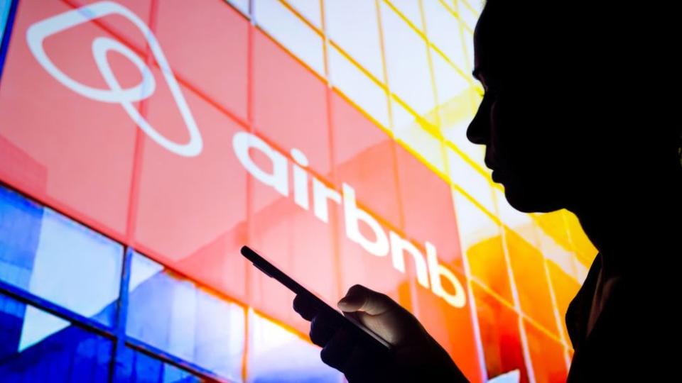 Sombra de una mujer sosteniendo un teléfono y con el logo de Airbnb detrás.